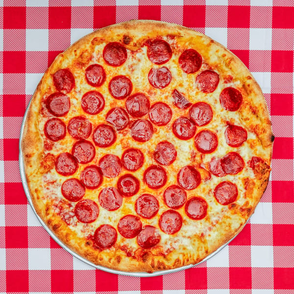 A whole pepperoni pizza on a metal tray, centered on a red and white checkered tablecloth.