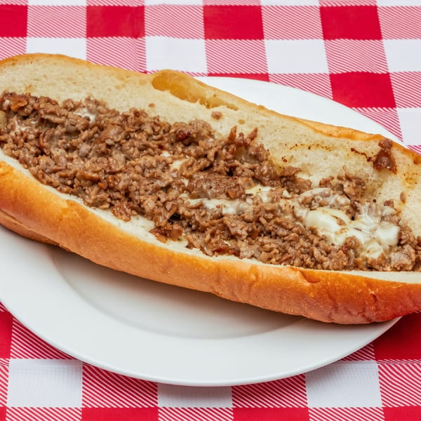 A cheesesteak with chopped steak and melted cheese on a fresh roll on a white plate over a red and white checkered tablecloth.