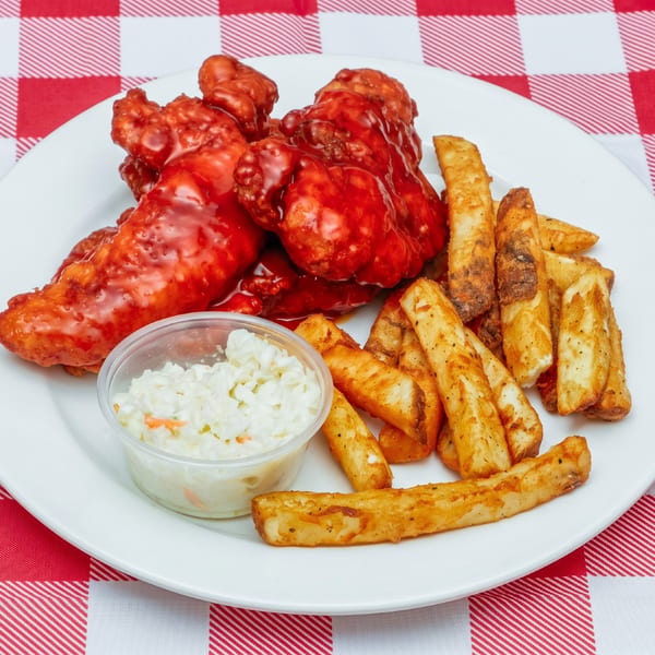 Buffalo chicken tenders with seasoned fries and a side of coleslaw on a white plate over a red and white checkered tablecloth.