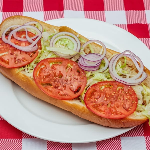 A hoagie topped with sliced tomatoes, shredded lettuce, and red onion rings on a white plate over a red and white checkered tablecloth.