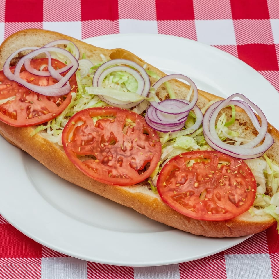 A hoagie topped with sliced tomatoes, shredded lettuce, and red onion rings on a white plate over a red and white checkered tablecloth.