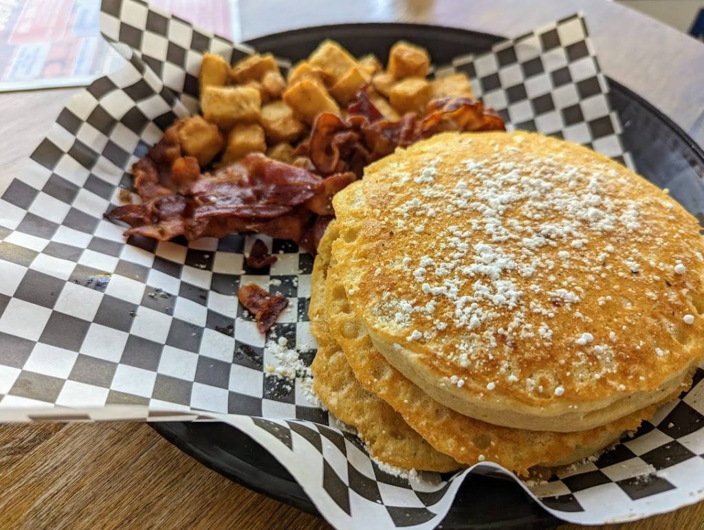 A stack of pancakes on a plate with bacon and tater tots.